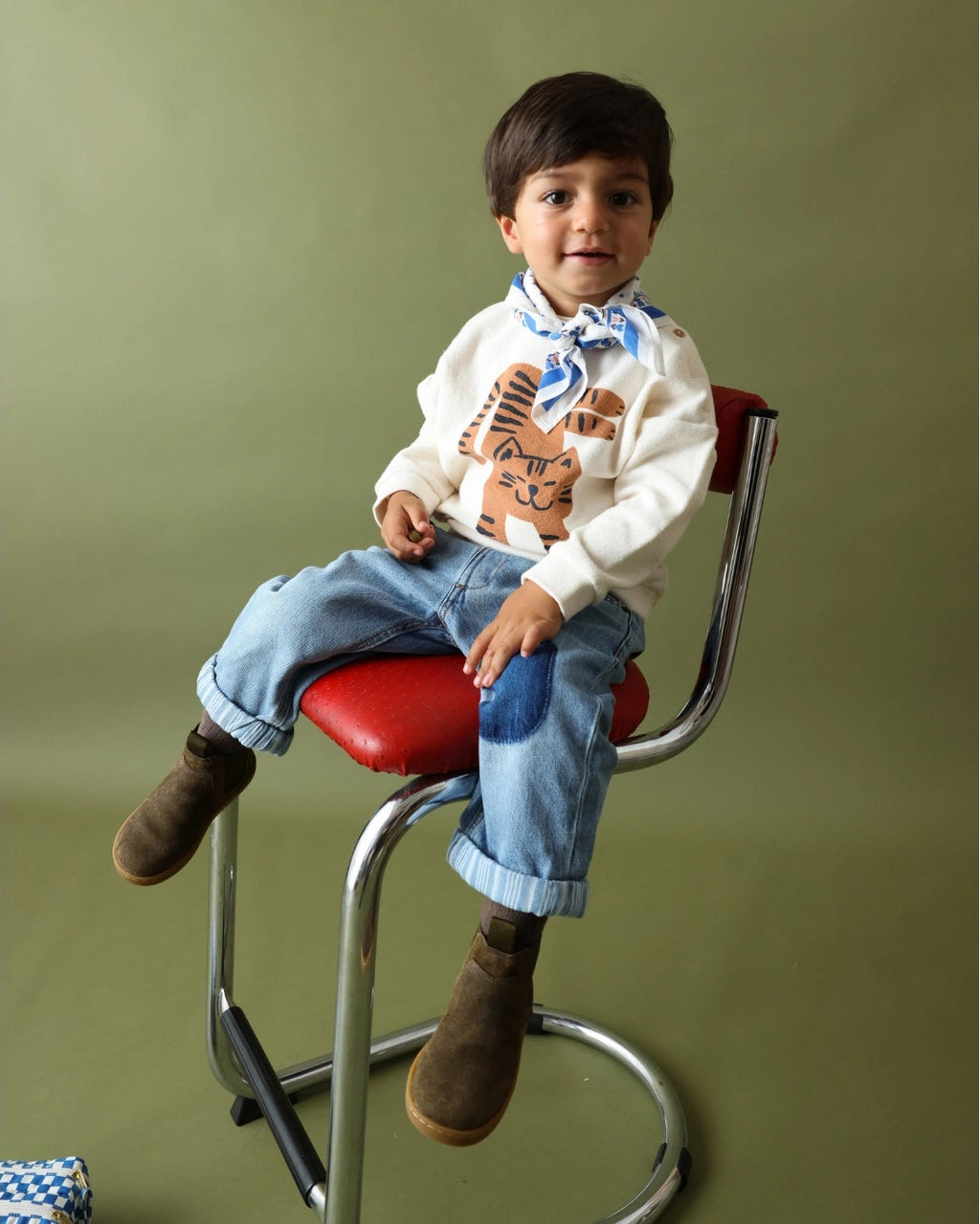 A young boy wearing a white long-sleeve shirt with a brown bear graphic, blue jeans, and brown boots sits on a red and chrome stool against an olive green background.