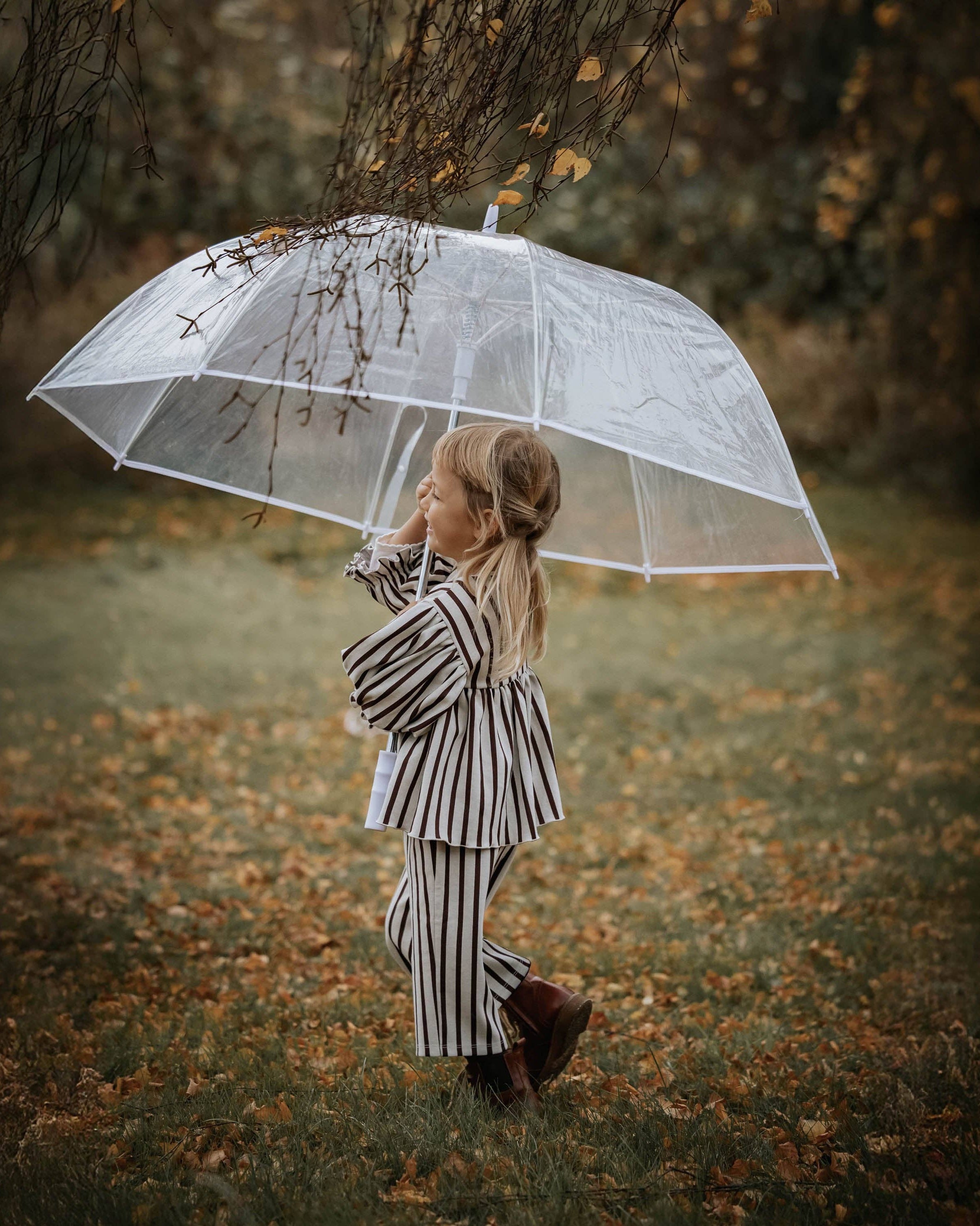 A young girl in a black and white striped pajama set walks under a clear transparent umbrella on a grassy path covered with fallen leaves, with trees in the background.
