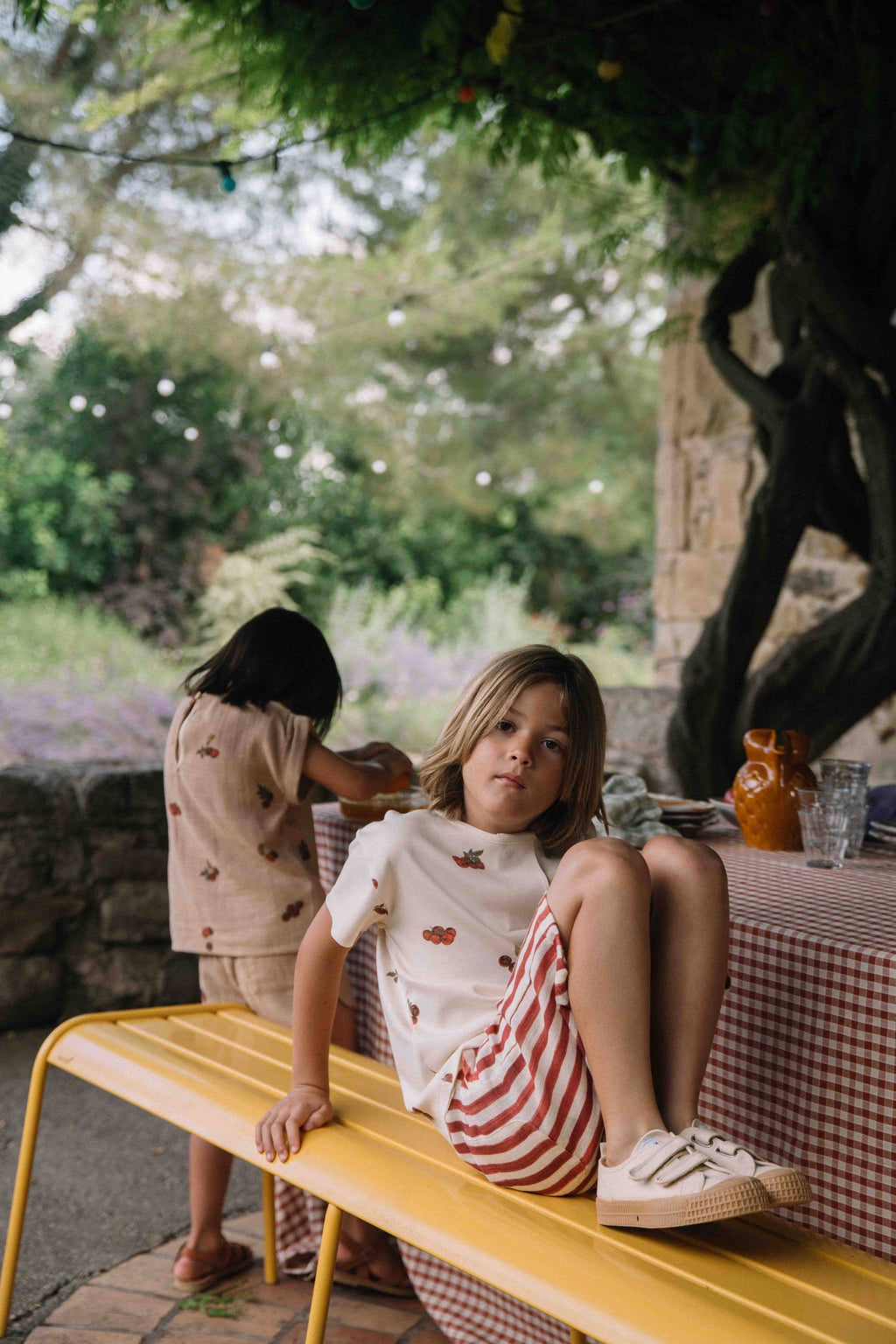Two children sitting on a yellow bench outdoors with a table and garden in the background.
