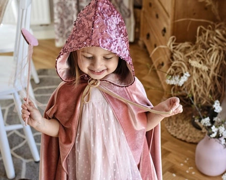 A young child wearing a pink hooded cape with a sequined top and a light pink dress, holding a wand in a cozy indoor room.