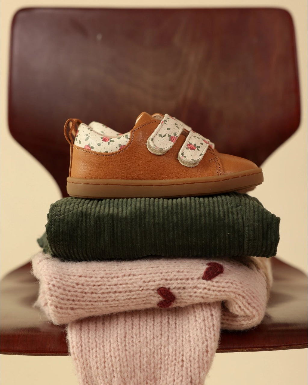A pair of tan slip-on shoes with floral-patterned soles stacked on top of folded knit socks in dark green and pale pink, placed on a wooden chair against a beige background.