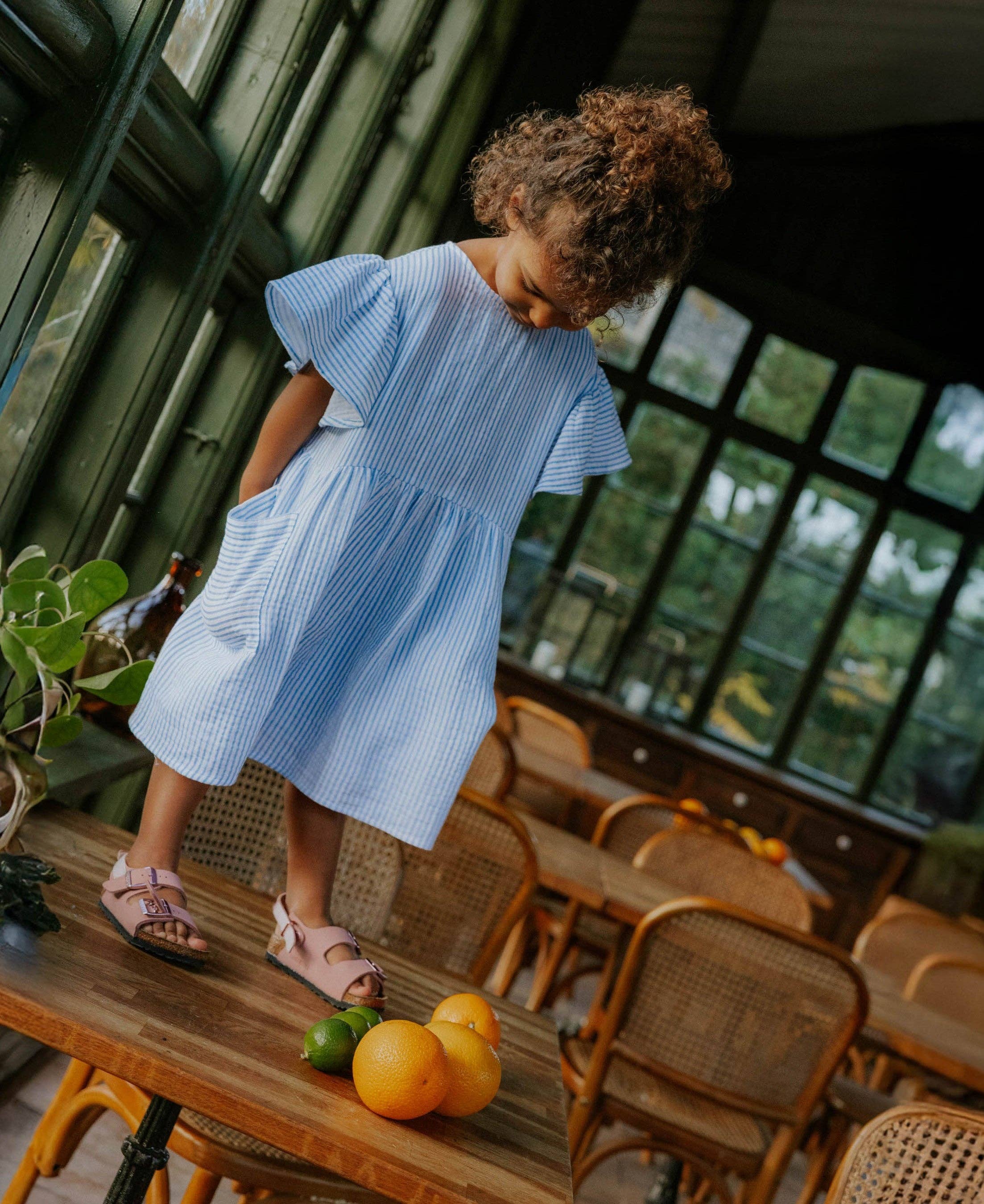 A young child with curly brown hair wearing a light blue and white vertically striped dress with flutter sleeves and side pockets, standing on a wooden table in a rustic cafe setting.