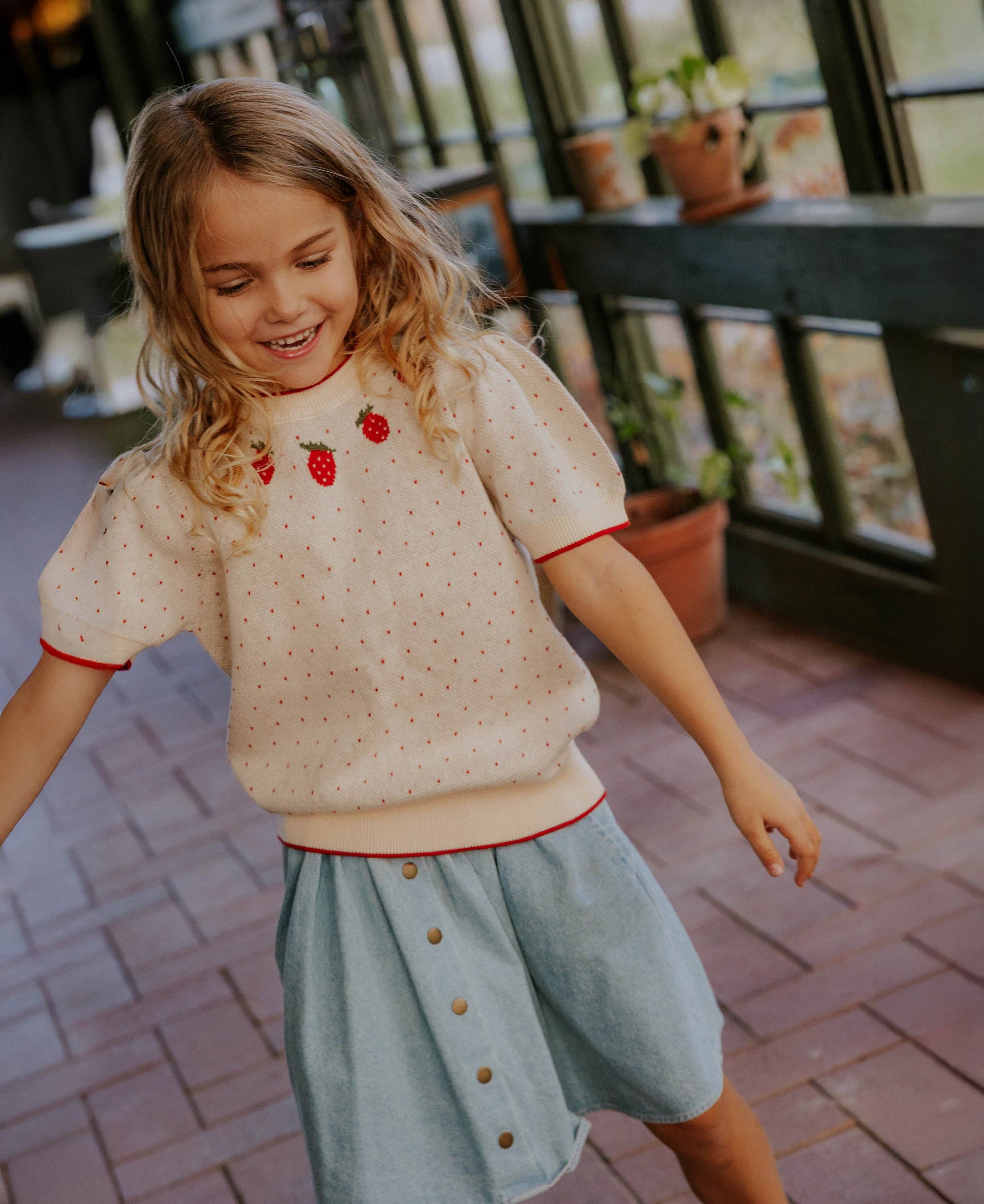 Young girl in a light-colored top with red accents and a light blue skirt outdoors.