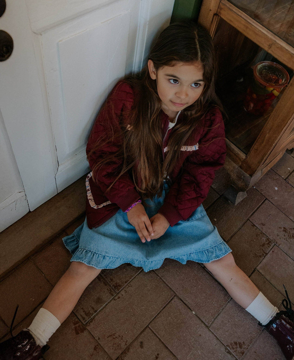A young girl with long brown hair sits on a brick floor in a rustic room, wearing a burgundy quilted jacket over a white top and a light blue denim skirt.