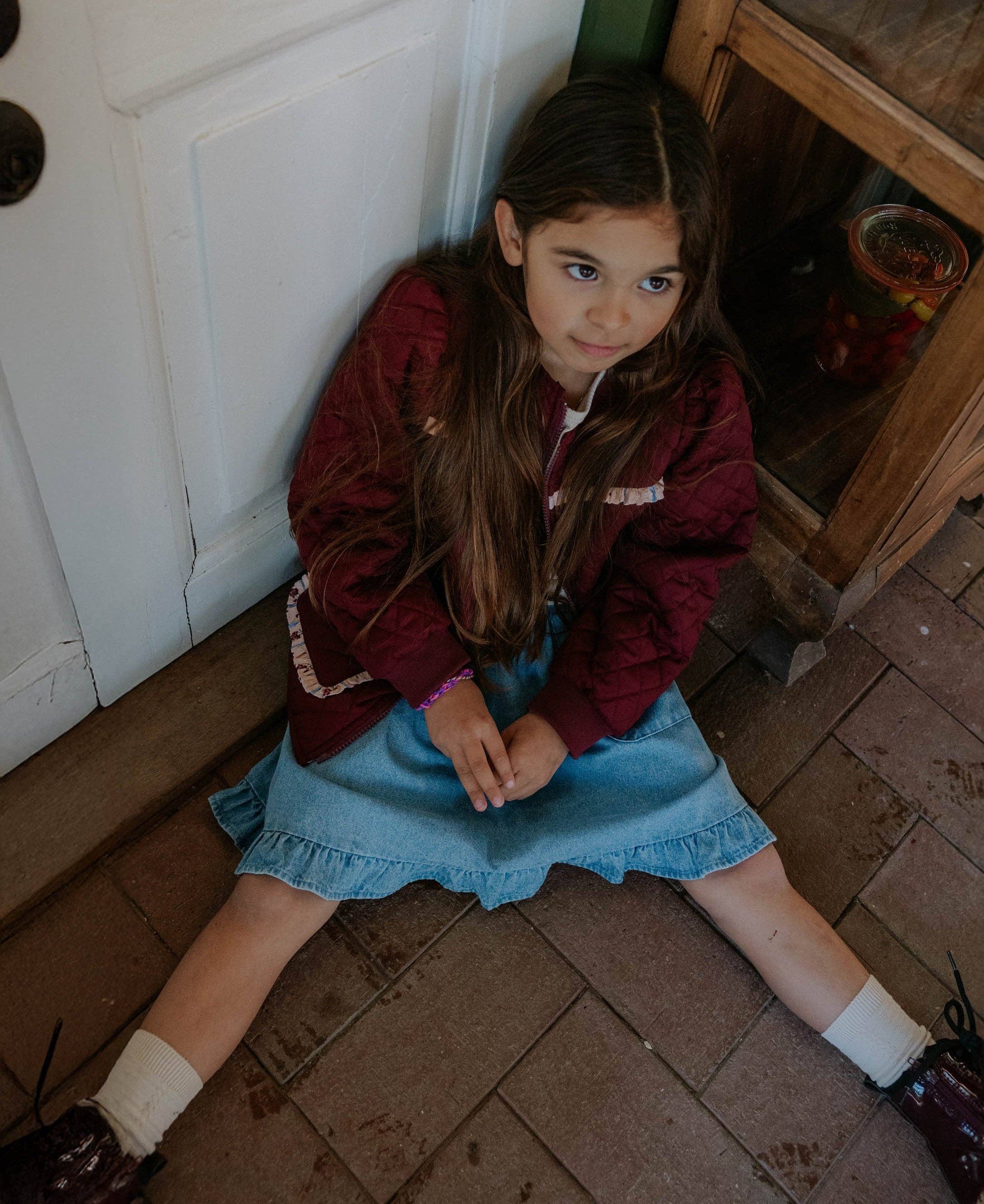 A young girl with long brown hair sits on a brick floor in a rustic room, wearing a burgundy quilted jacket over a white top and a light blue denim skirt.