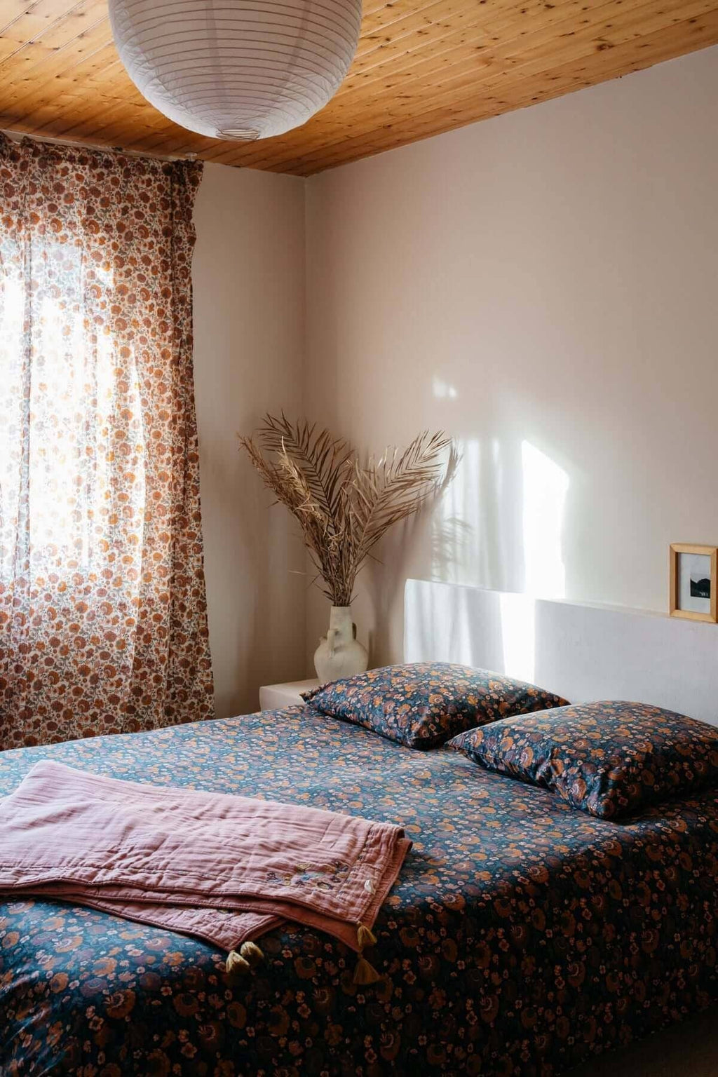 Bedroom with floral bedding, curtains, and a vase on a shelf.