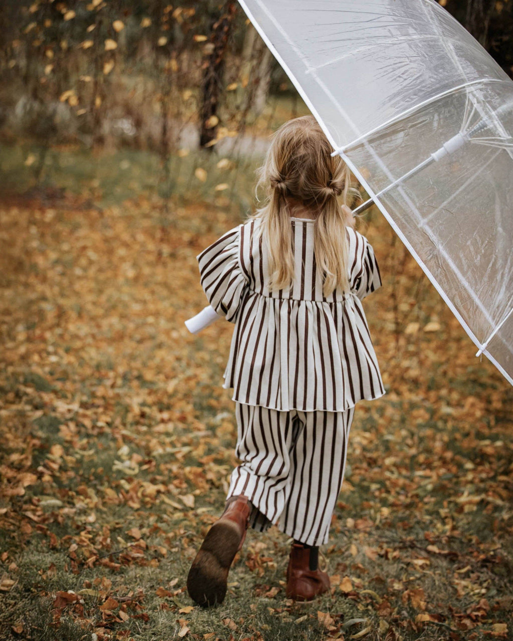 A young girl with blonde hair in a ponytail, seen from behind, wearing a black and white vertically striped two-piece outfit and walking in a leaf-covered outdoor path while holding a clear plastic umbrella.
