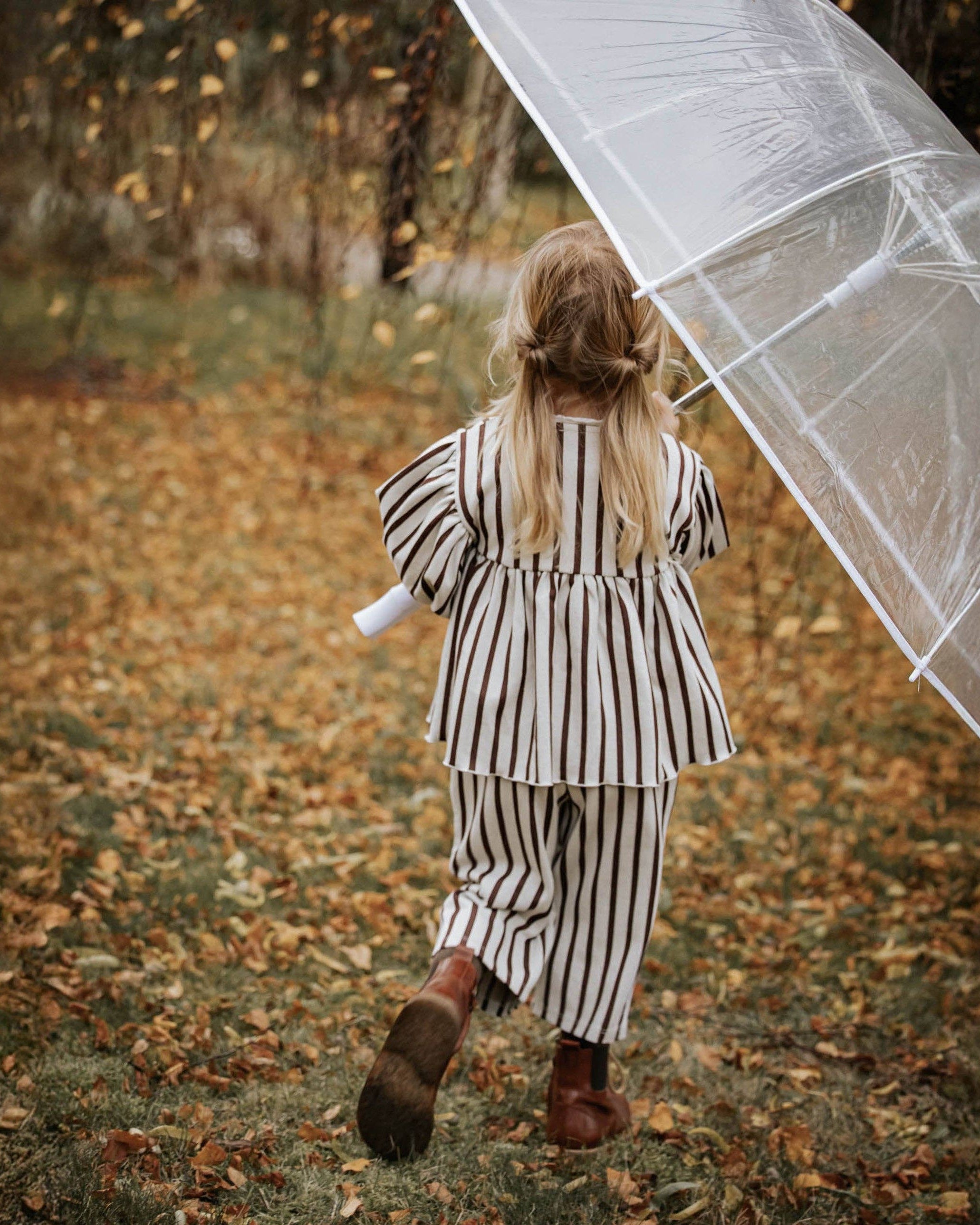 A young girl with blonde hair in a ponytail, seen from behind, wearing a black and white vertically striped two-piece outfit and walking in a leaf-covered outdoor path while holding a clear plastic umbrella.