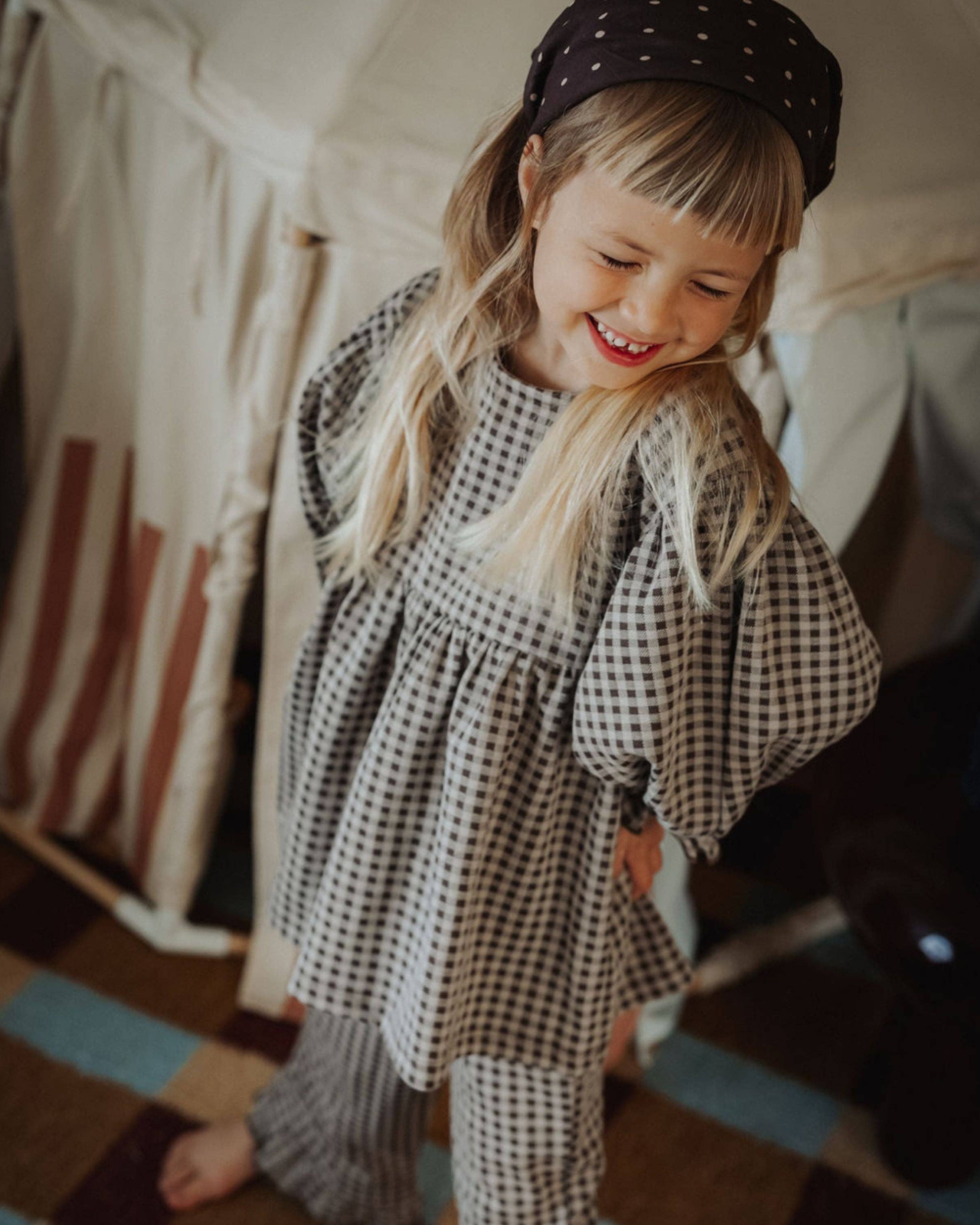 Child wearing a checkered outfit and polka dot hat inside a tent-like structure.