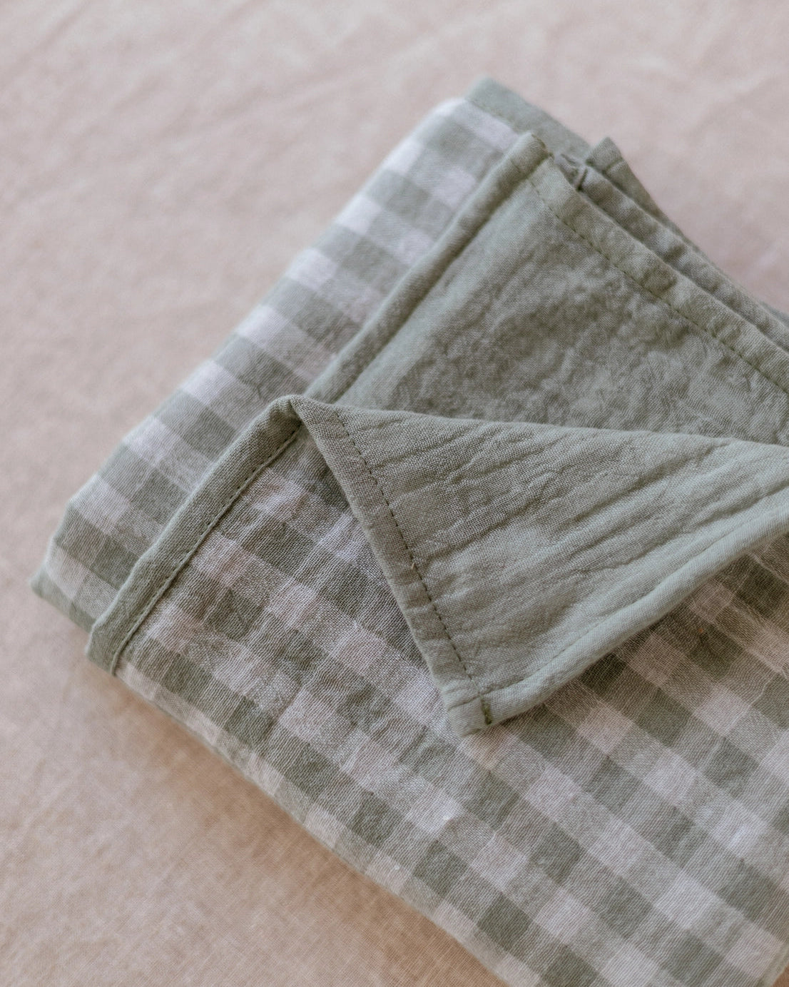 A folded sage green and off-white gingham check cloth with a soft, textured cotton-linen feel, displayed on a neutral beige fabric background.