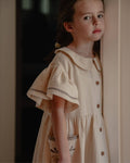 Young girl wearing a beige dress with embroidery, standing indoors.