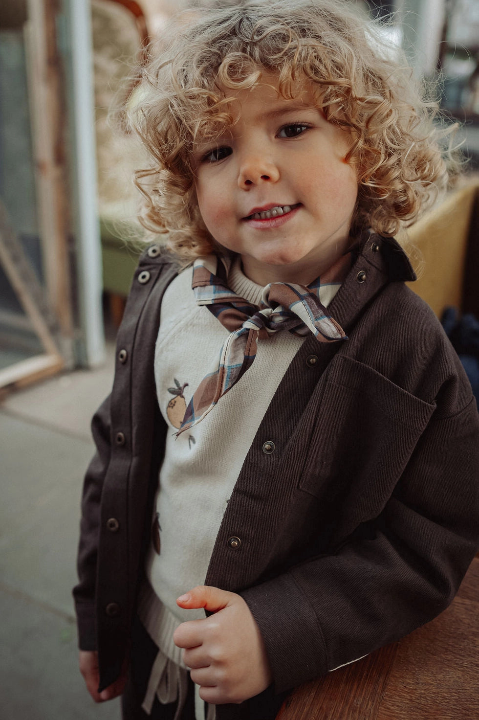A young child with curly blonde hair wearing a dark brown button-up jacket over a cream sweater with a plaid necktie, photographed in a softly blurred indoor setting.