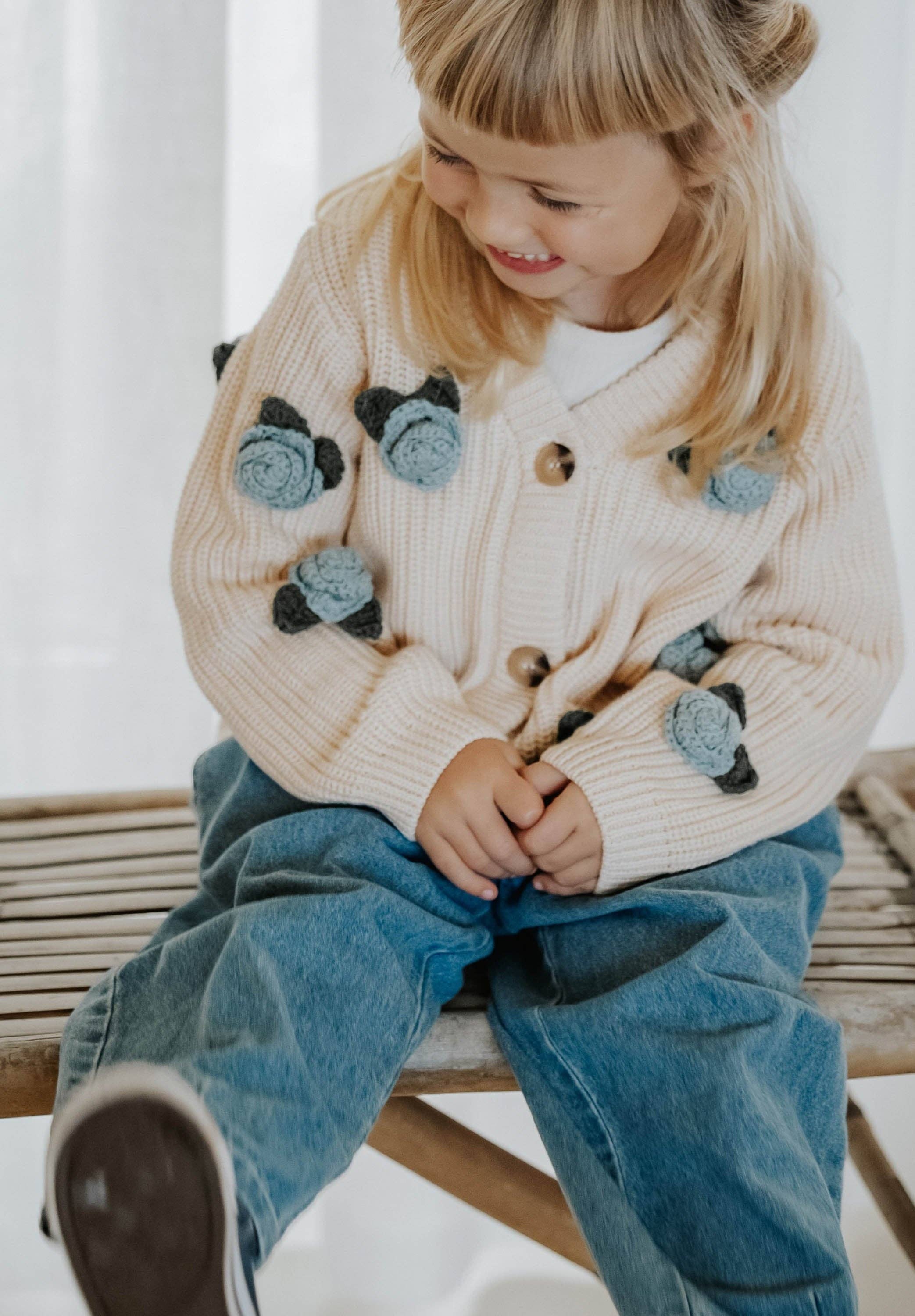Child wearing a beige cardigan with cat appliqués and blue jeans sitting on a wooden bench.