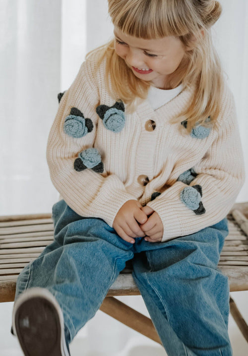 Child wearing a beige cardigan with cat appliqués and blue jeans sitting on a wooden bench.