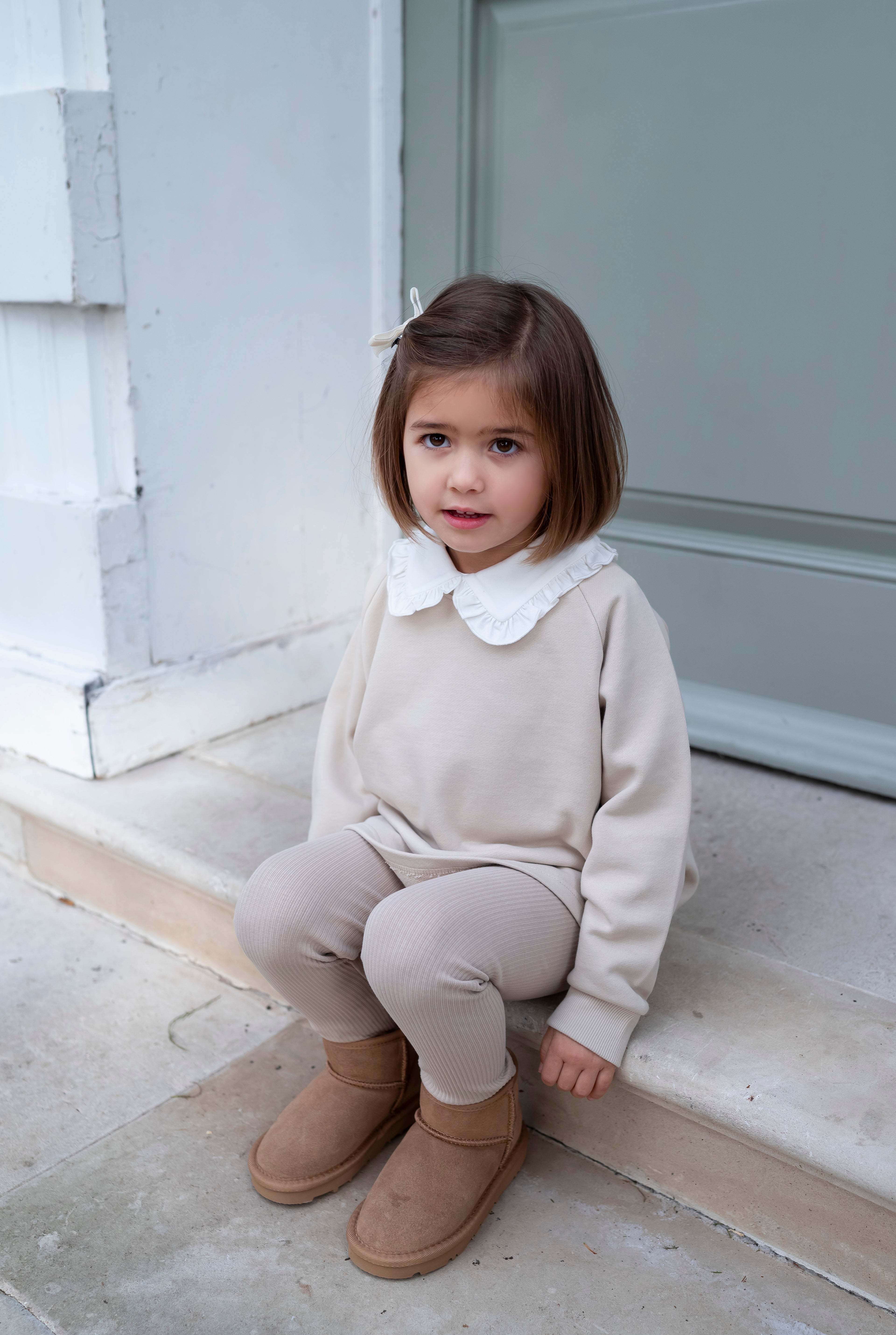 Young girl sitting on steps wearing a beige outfit with a white collar and brown boots.
