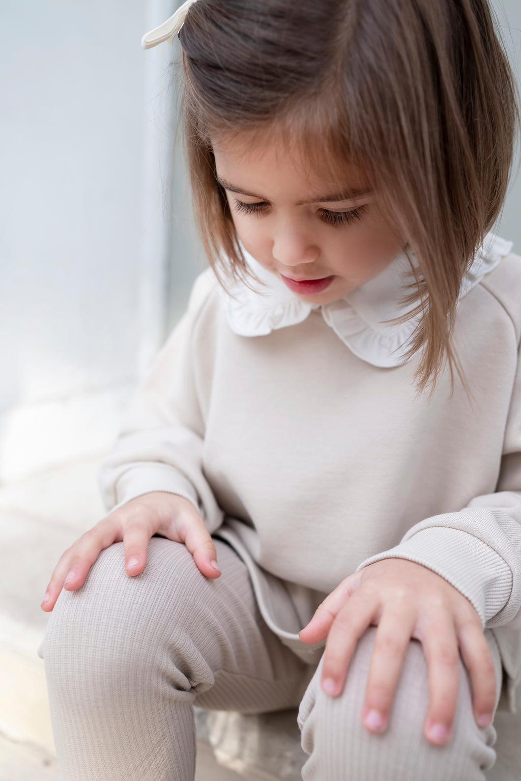 Young girl in a beige outfit with a white collar sitting on the floor.