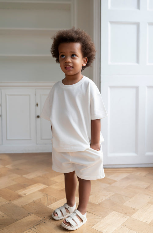 Child wearing a white outfit standing in a room with wooden flooring and white walls.