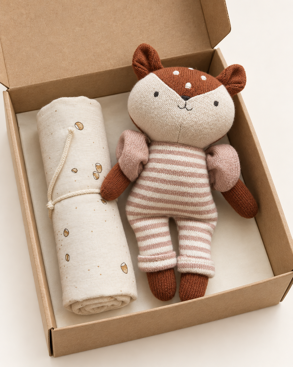 A soft fabric toy deer doll wearing pink and cream striped clothing, displayed next to a rolled cream blanket with small gold leaf prints inside an open brown cardboard gift box.