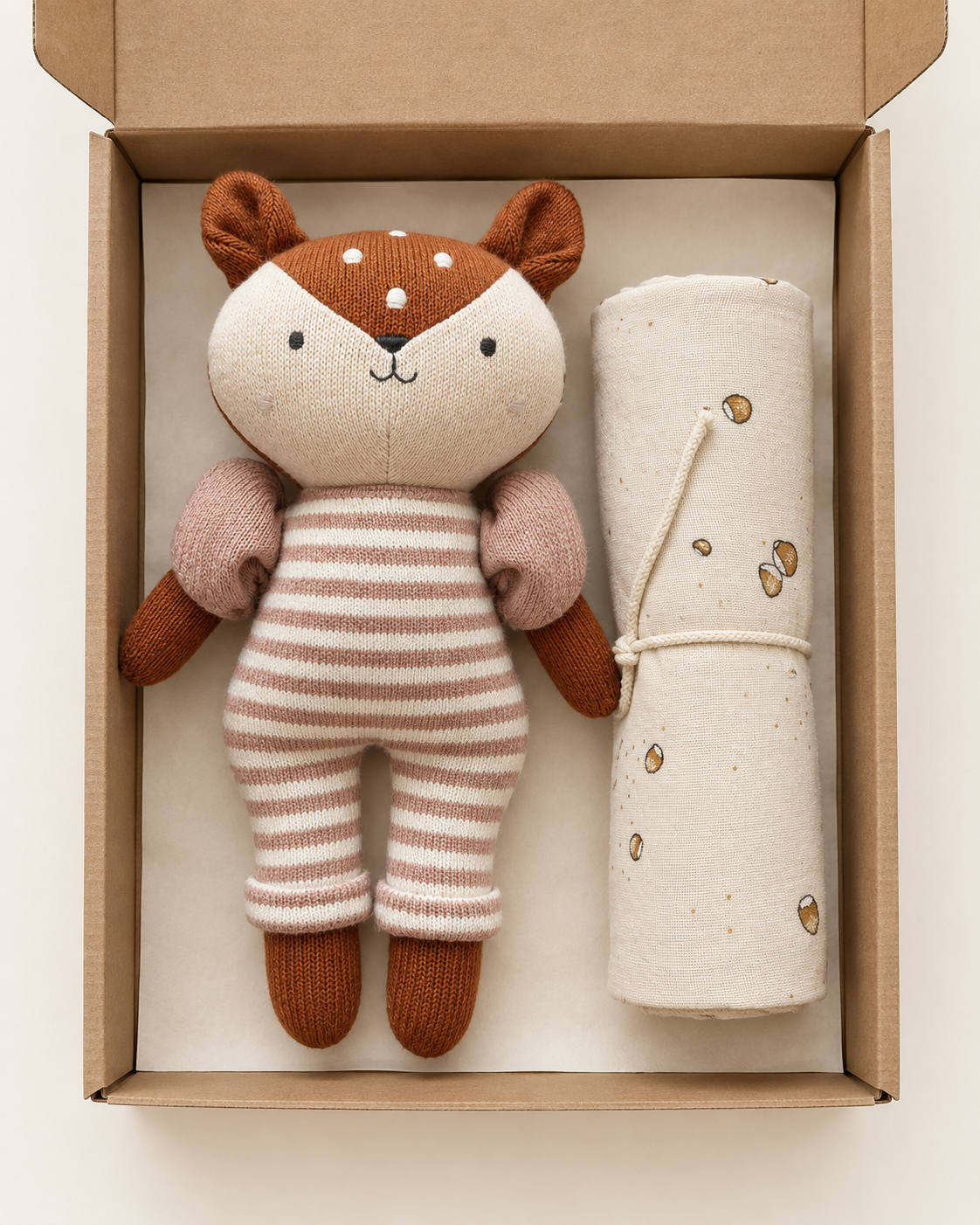 A plush stuffed animal toy deer, brown ears and feet, and pink-and-white striped body, displayed inside an open brown cardboard box next to a rolled cream fabric item with small tan spots.