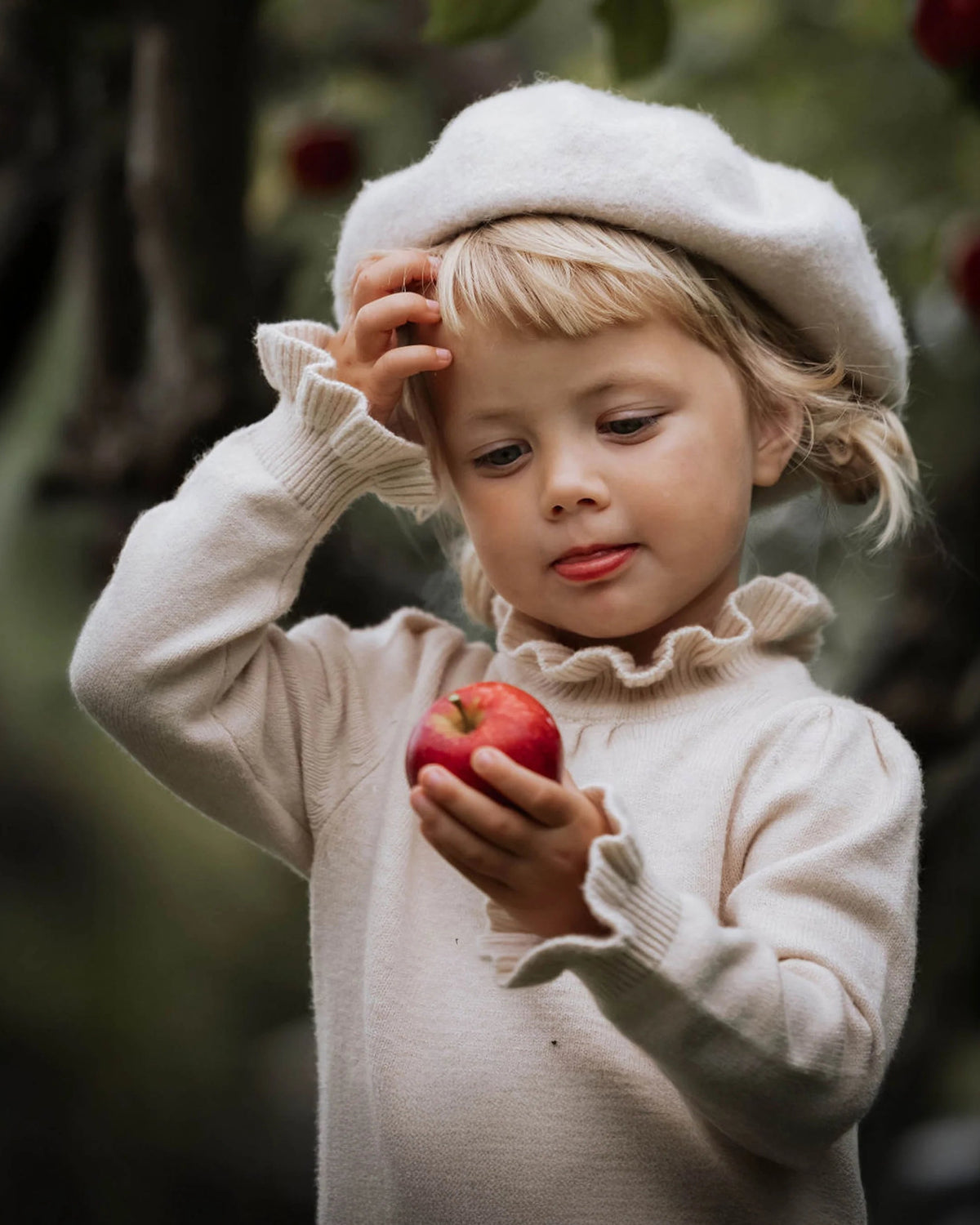 A young blonde girl wearing a cream-colored knit sweater and matching beret hat, holding a red apple in an outdoor garden setting.