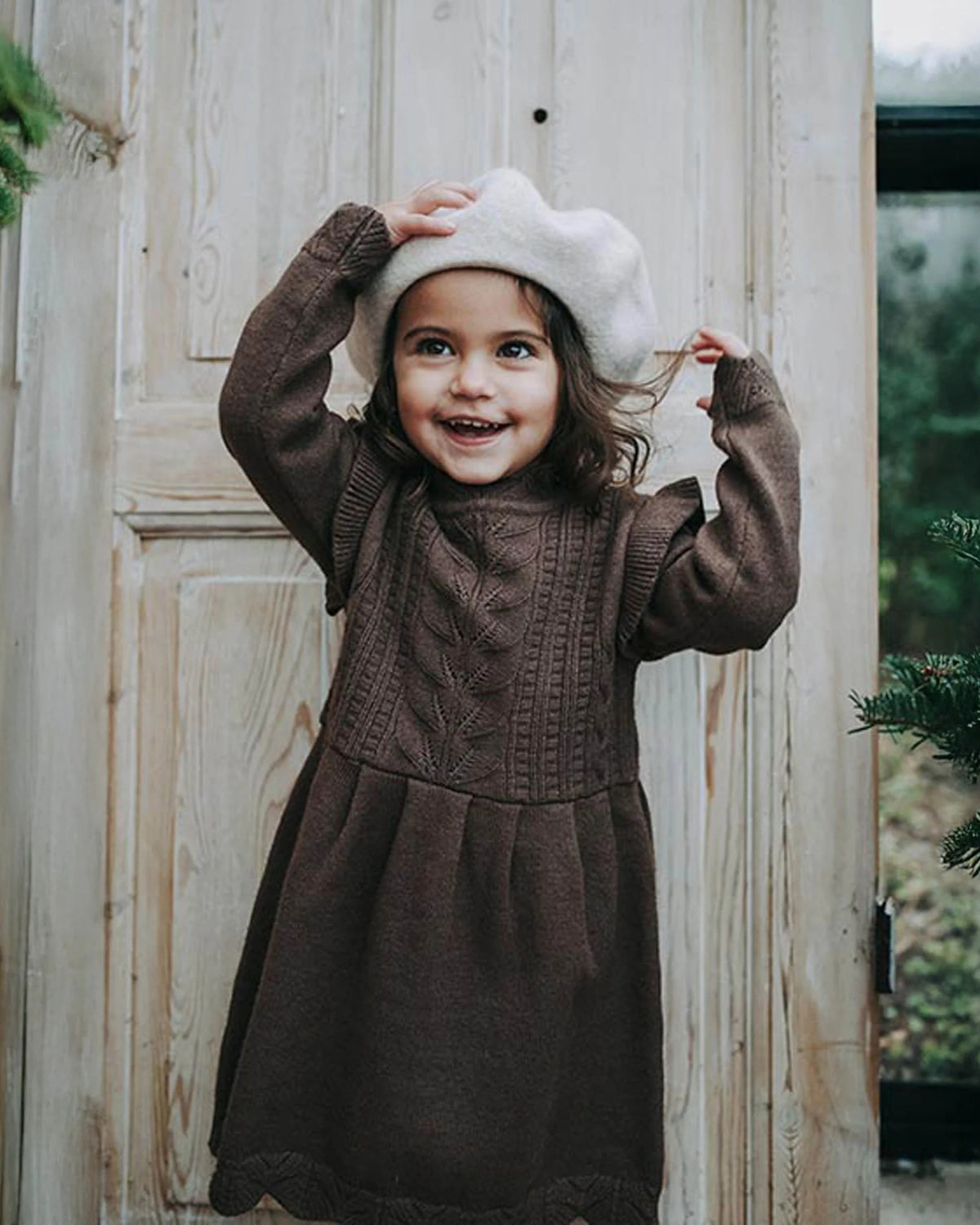 A young girl smiling and posing in front of a rustic wooden door, wearing a dark brown cable-knit dress and a white beret.
