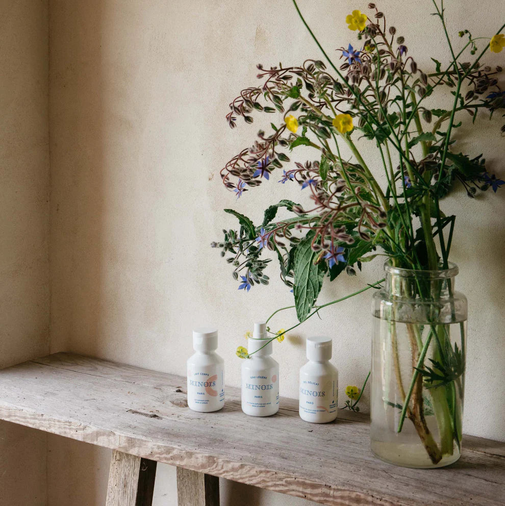 Three white bottles on a wooden bench with a vase of wildflowers against a beige wall.