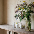 Three white bottles on a wooden bench with a vase of wildflowers against a beige wall.