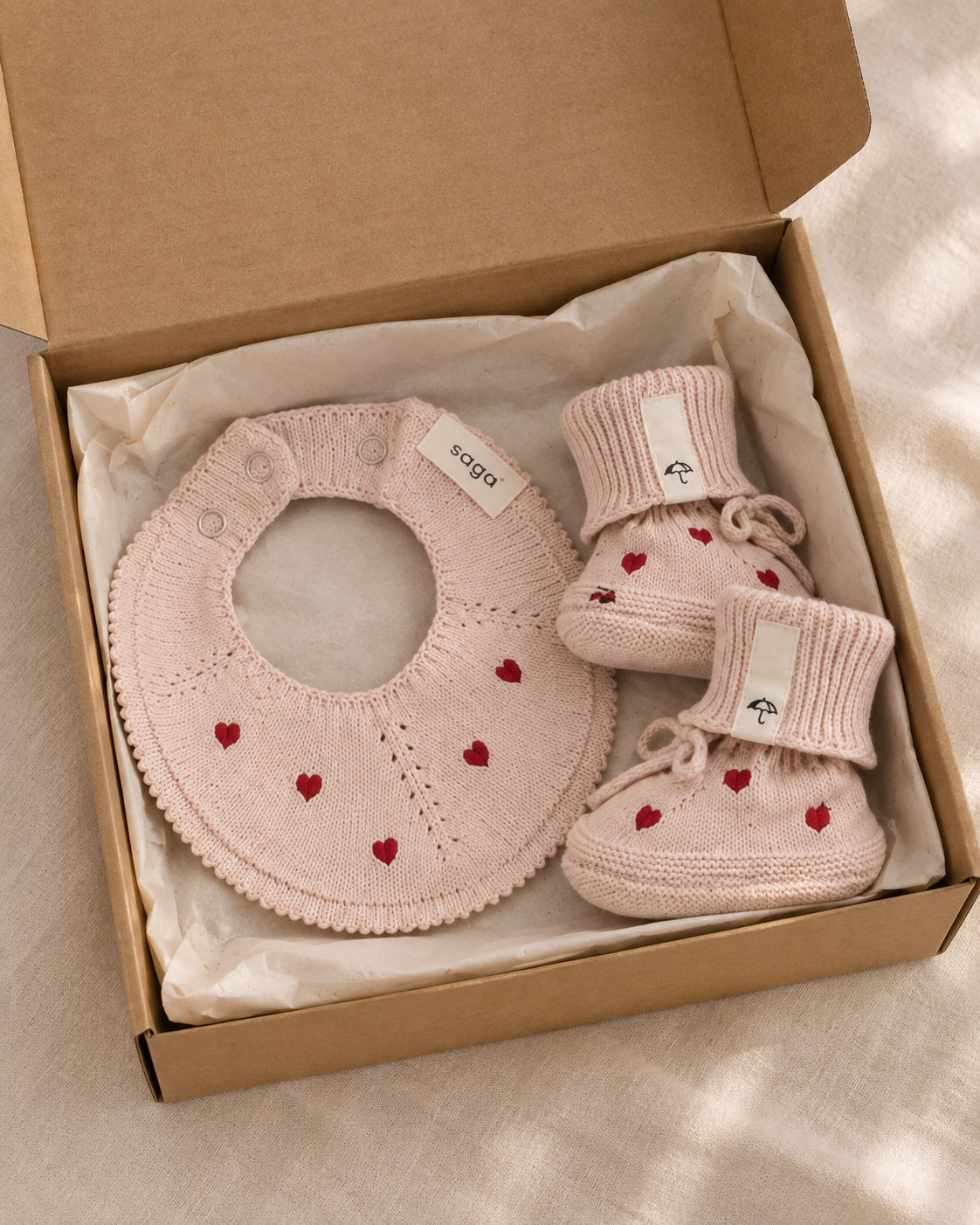 A baby gift set including a light pink knit bib and matching booties with small red heart embroidery, presented in an open brown cardboard box lined with white tissue paper.