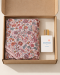 A perfume bottle labeled MINOIS Paris next to a folded floral scarf, presented inside an open brown cardboard gift box on a light beige background.