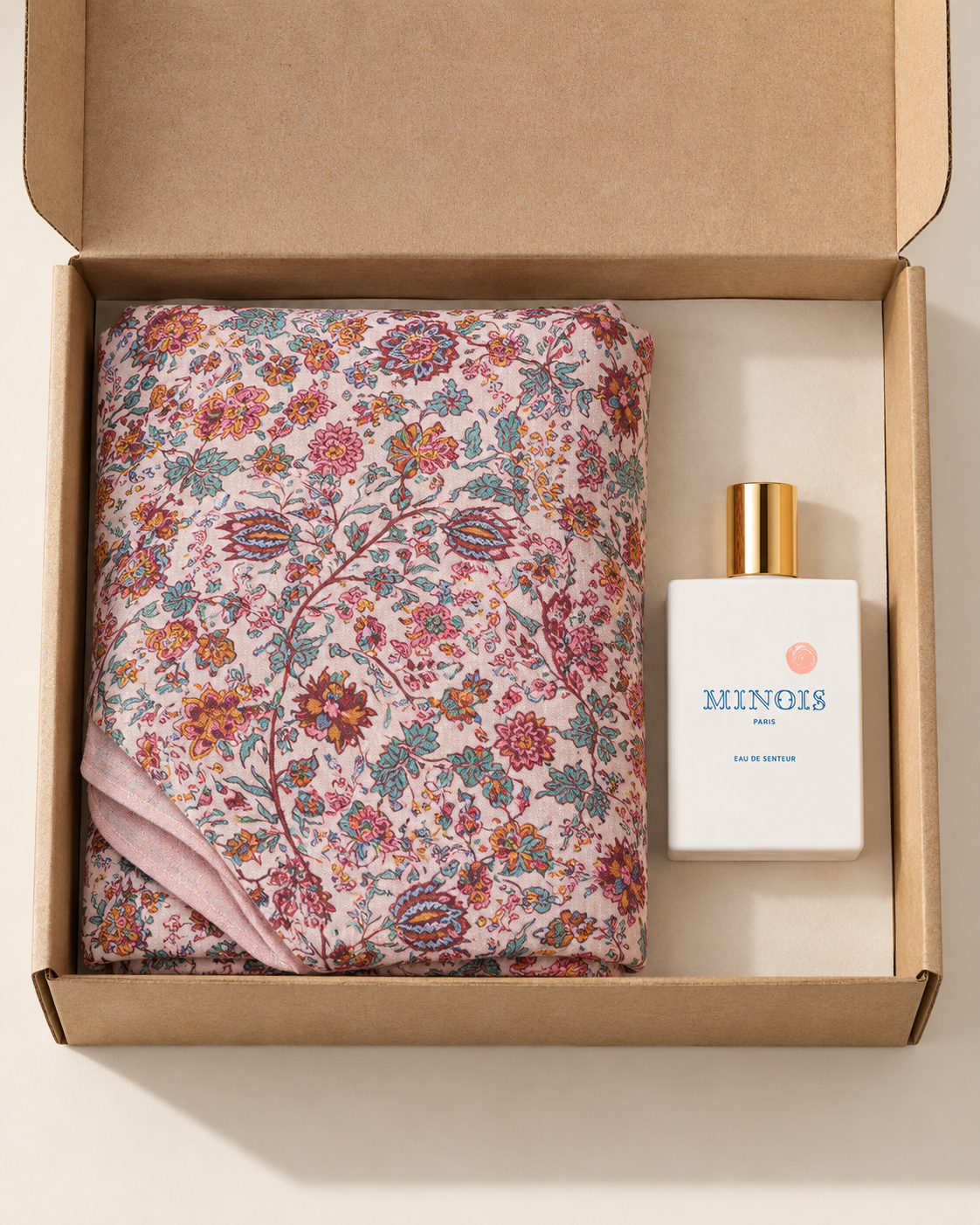 A perfume bottle labeled MINOIS Paris next to a folded floral scarf, presented inside an open brown cardboard gift box on a light beige background.