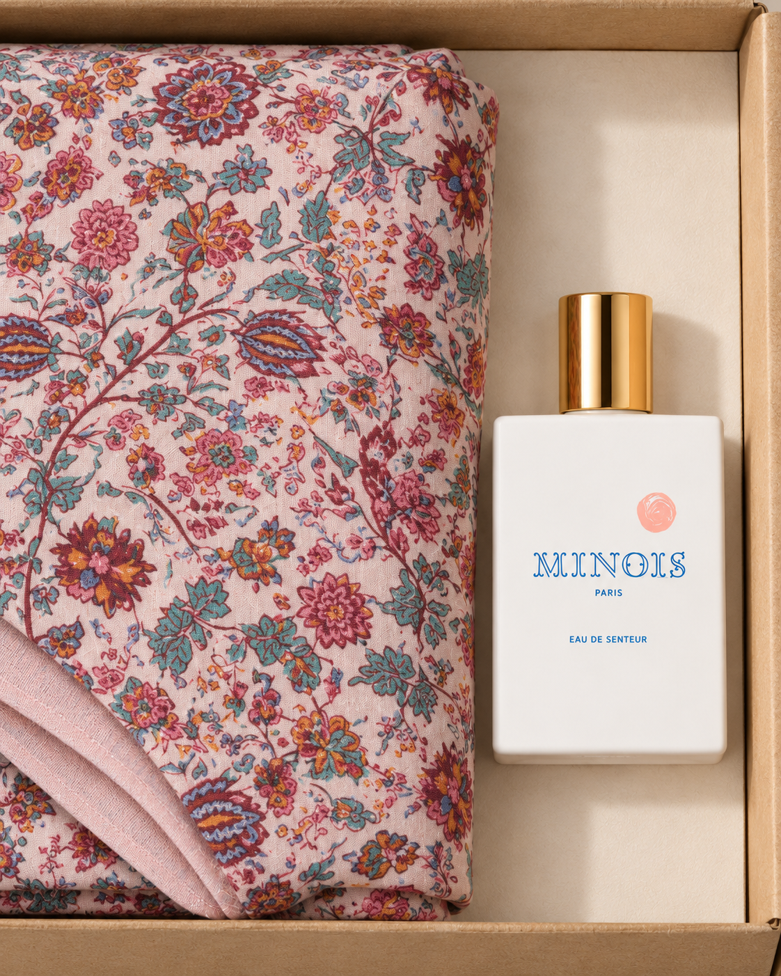 A perfume bottle labeled MINOIS PARIS next to a folded pink floral textile inside a light brown gift box.