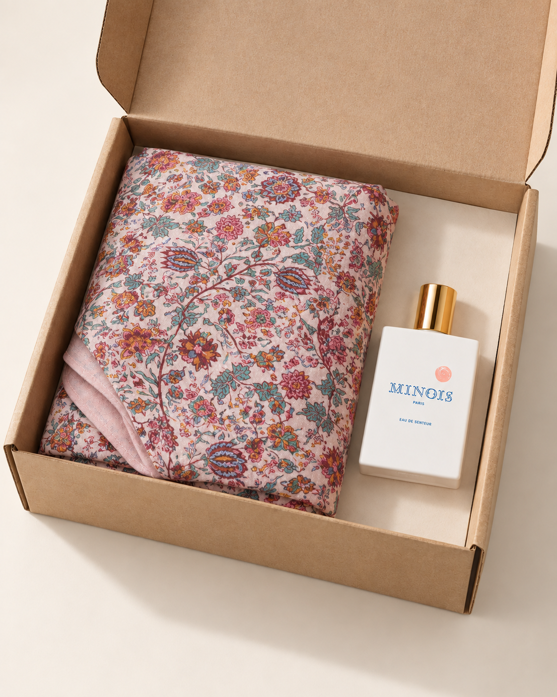 A gift set in a brown kraft box featuring a folded pink floral scarf and a white MINOIS Paris cologne bottle with a gold cap.