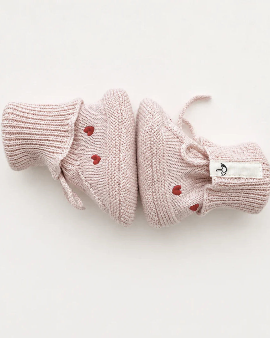 A pair of pale pink knit baby booties with small red heart details and white laces, laid flat on a clean white background.