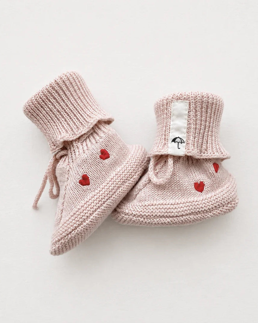 A pair of light pink knit baby booties with small red embroidered hearts and drawstring ankle ties, displayed on a clean off-white background.