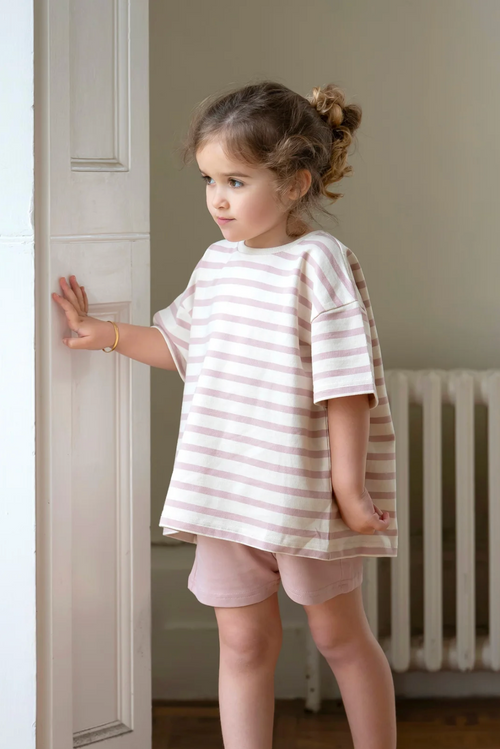 Young girl in a striped shirt and shorts standing in a room with a radiator.