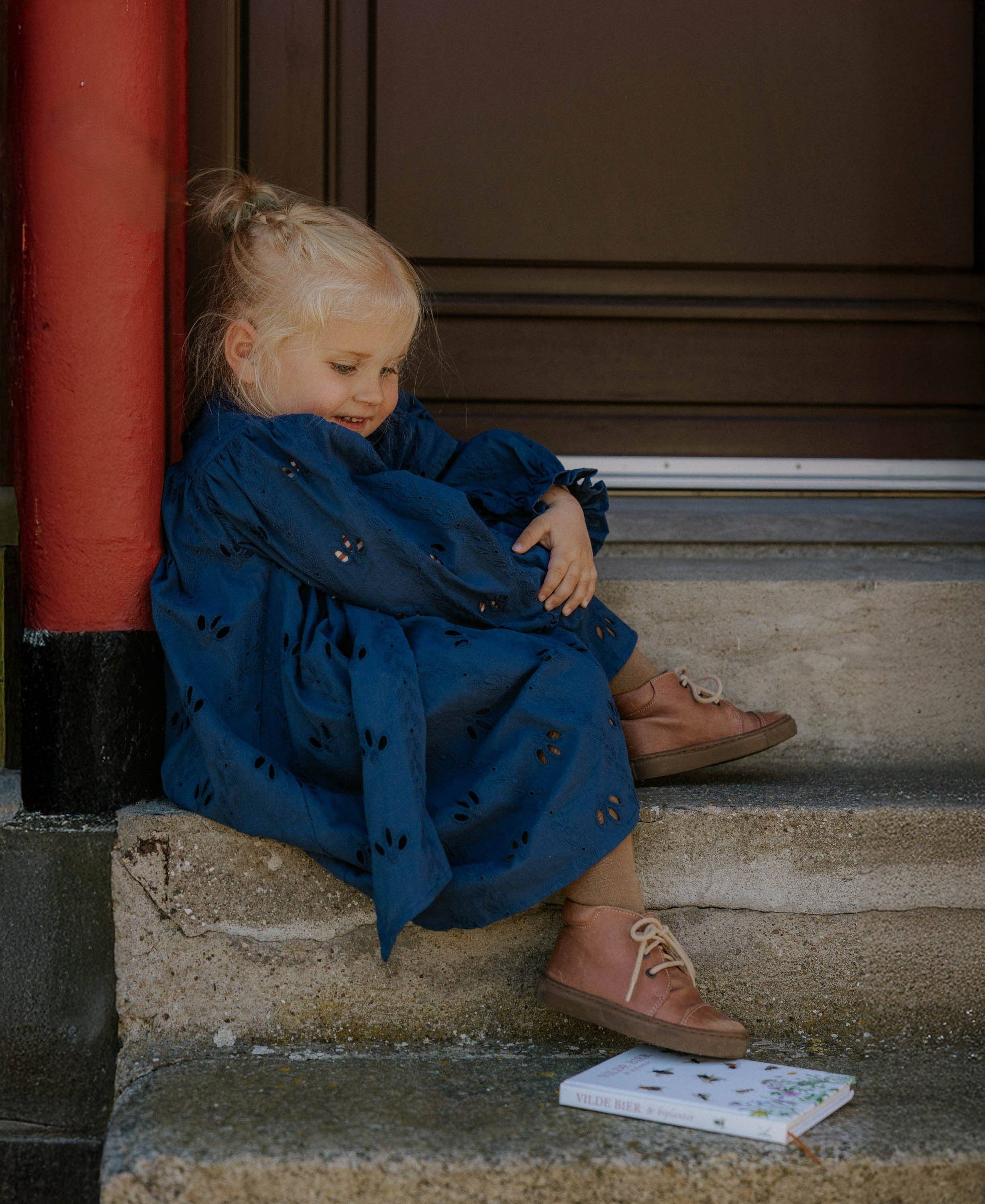 Child in a blue dress sitting on steps with a book beside them