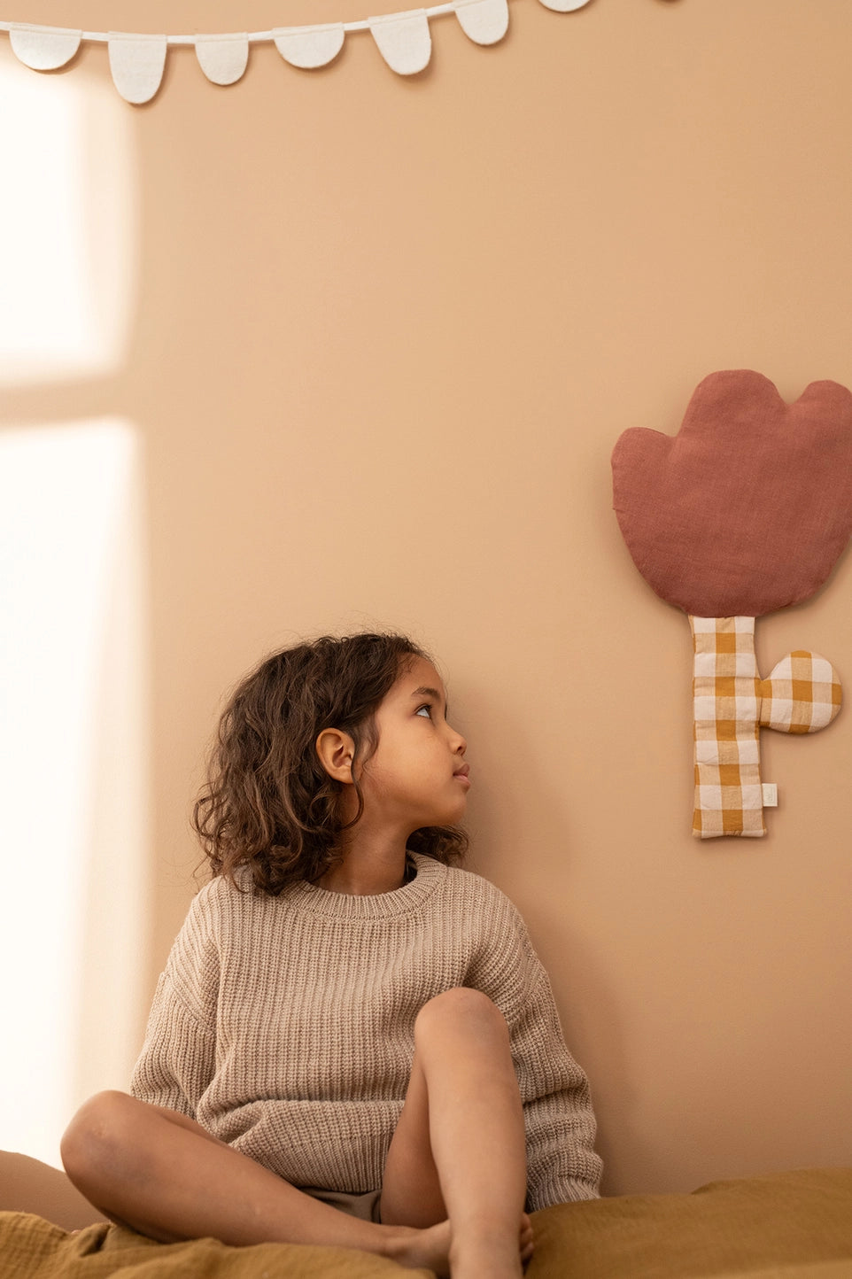 A young girl with curly brown hair sits on a mustard yellow bed, looking up at a peach-colored wall decorated with a brown flower-shaped wall hanging and a white scalloped banner.