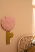 A pink flower-shaped cushion with a mustard yellow stem and leaf, mounted on a warm beige wall next to a rattan headboard.