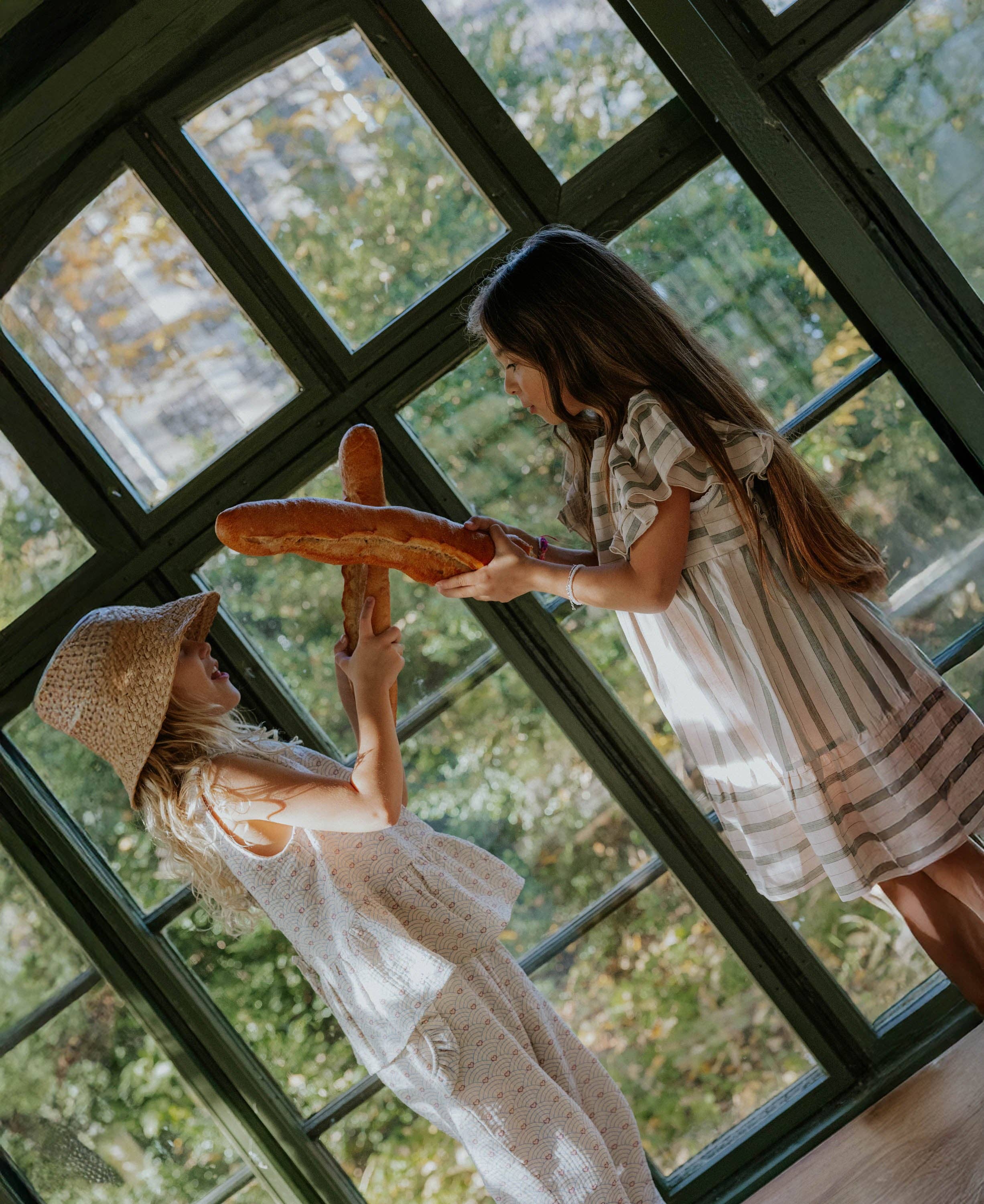 Two girls holding baguettes in front of a large window with a view of greenery.