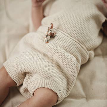 A close-up of a baby wearing a cream-colored, textured knit romper with a small embroidered butterfly detail on the chest, lying on a soft beige blanket.