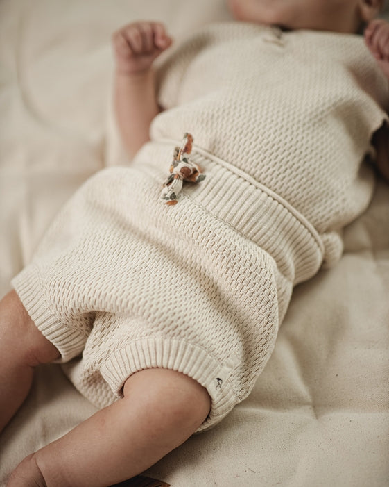 A close-up of a baby lying on a soft beige blanket, wearing a cream-colored waffle-knit romper with a small decorative animal applique at the waist.