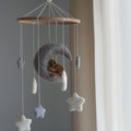 A felt baby mobile with a wooden hoop, featuring a gray crescent moon cradling a small brown teddy bear, surrounded by hanging white and blue-gray stars and clouds, photographed indoors near a window.