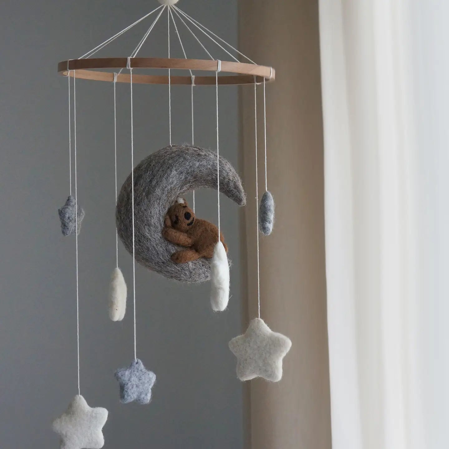A felt baby mobile with a wooden hoop, featuring a gray crescent moon cradling a small brown teddy bear, surrounded by hanging white and blue-gray stars and clouds, photographed indoors near a window.