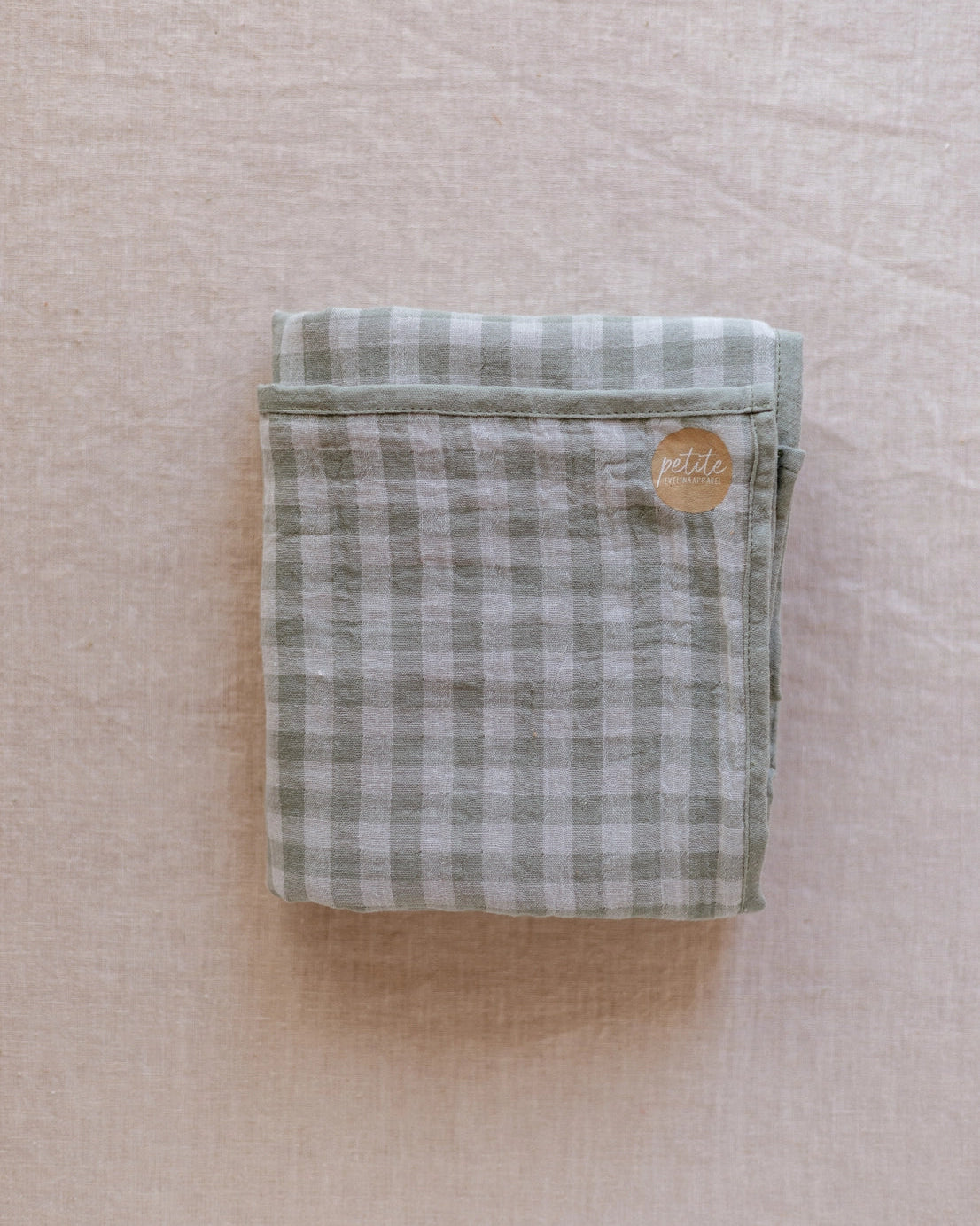 A folded cotton kitchen towel with a sage green and white gingham check pattern, featuring a small circular gold metal brand tag that reads 'pith'. The towel is laid flat on a textured, light beige linen fabric background.