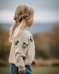 Child wearing a cream sweater with green buttons in a natural setting