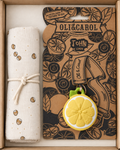 A gift set in a light brown box containing a rolled cream cotton blanket with a small brown and gold pattern and a cream rope tie, alongside a branded Oli & Carol fruit & veggie pouch card featuring lemon illustrations and a yellow lemon-shaped key ring charm.