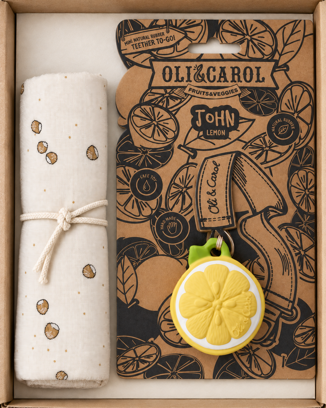 A gift set in a light brown box containing a rolled cream cotton blanket with a small brown and gold pattern and a cream rope tie, alongside a branded Oli & Carol fruit & veggie pouch card featuring lemon illustrations and a yellow lemon-shaped key ring charm.