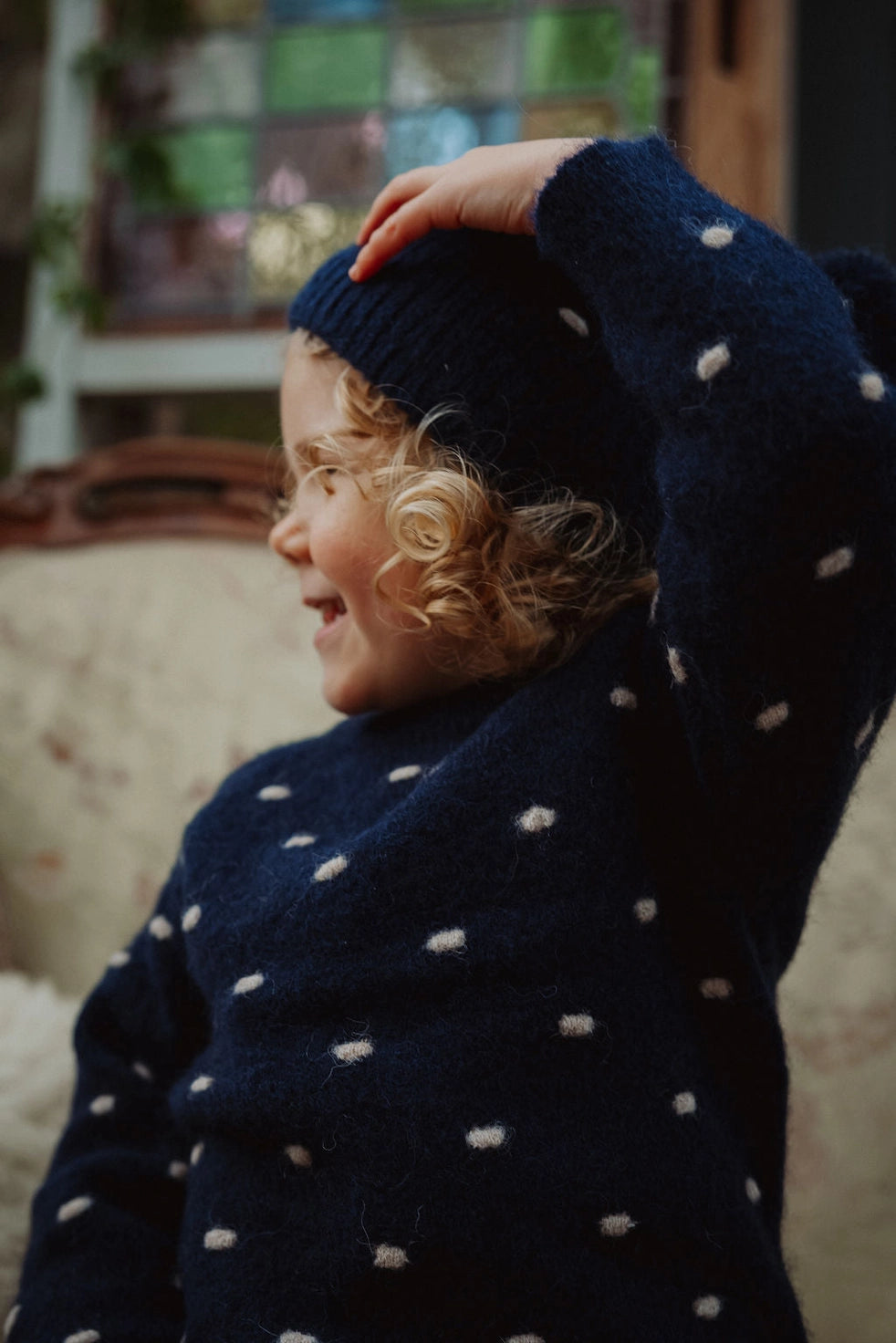 A child wearing a navy blue knit sweater and matching beanie with white polka dots, smiling and looking to the side in a cozy indoor setting.