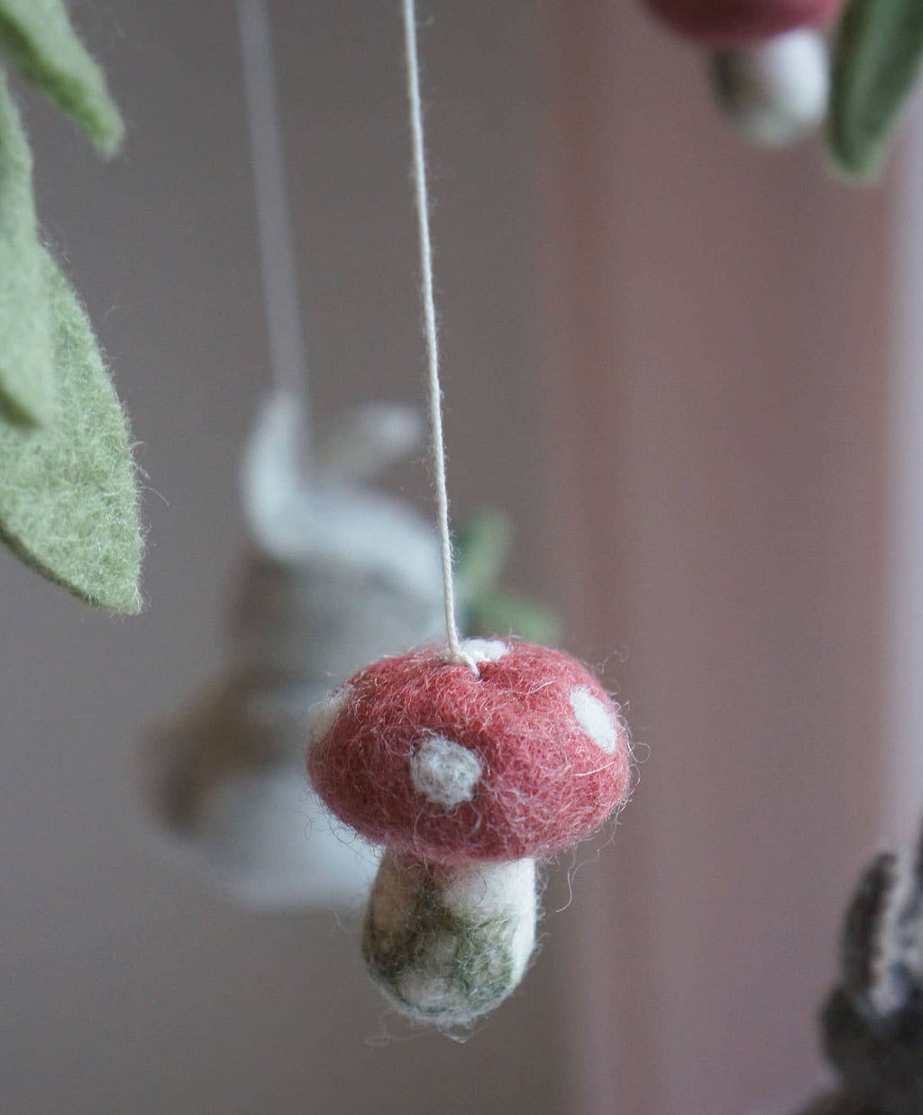 A close-up of a felted mushroom ornament hanging from a thin string, with a soft pink background and blurred green leaves.