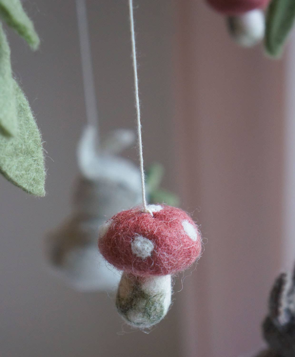 A close-up of a felted mushroom ornament hanging from a thin string, with a soft pink background and blurred green leaves.